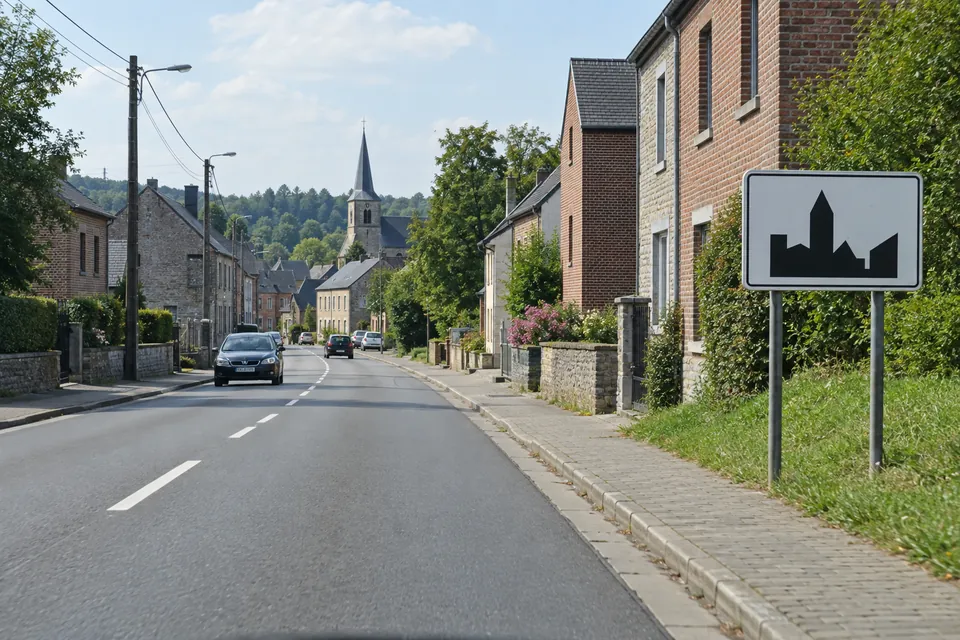 Entrée d’une agglomération wallonne, avec un panneau blanc représentant une silhouette de bâtiments, sans limitation chiffrée supplémentaire.