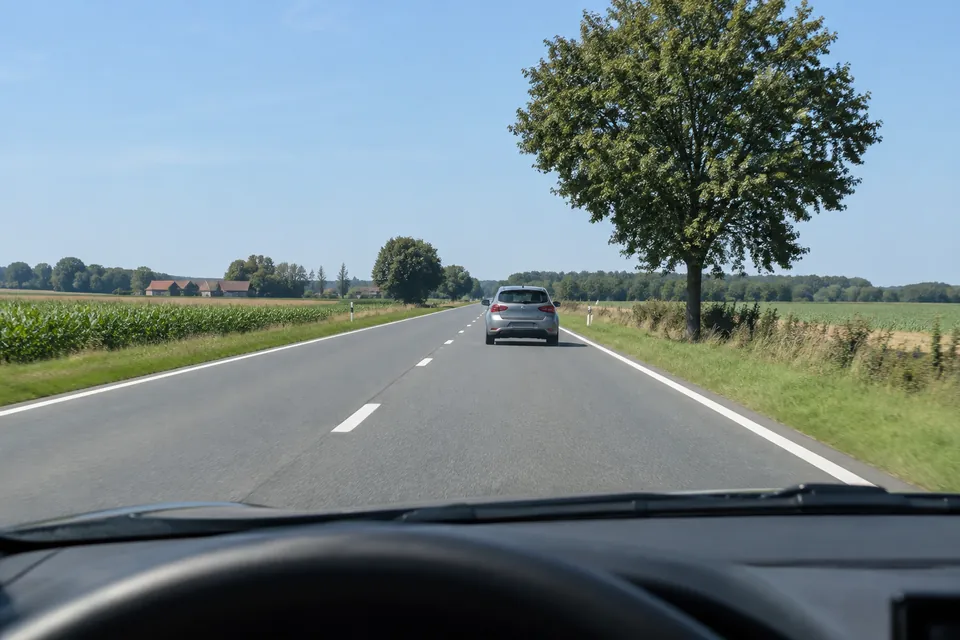 Voiture suivie sur une route sèche, avec un arbre en bordure et de la chaussée visible entre les véhicules.