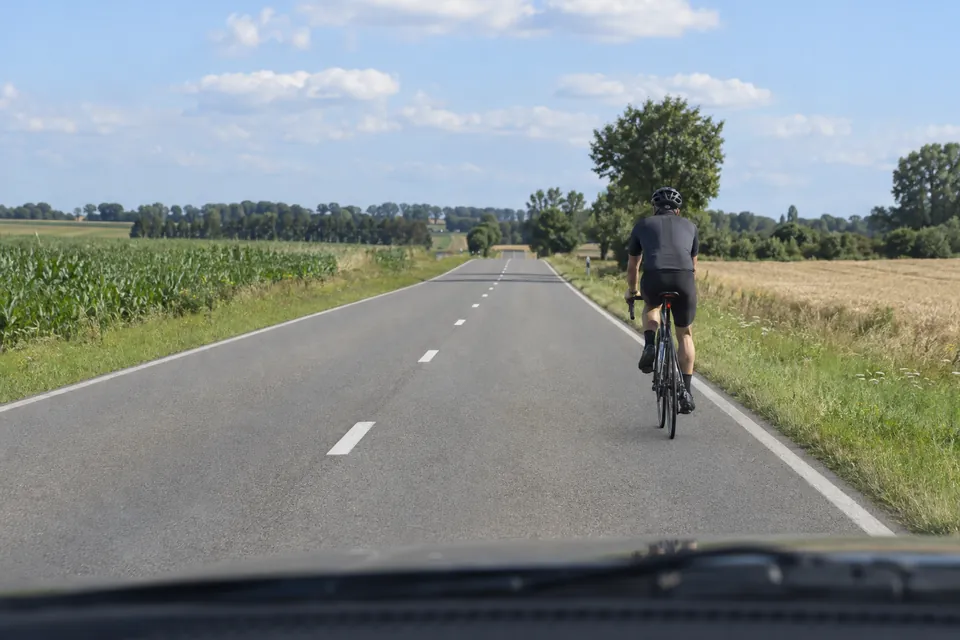Cycliste vu de dos sur le côté droit d’une route rurale entre des champs.