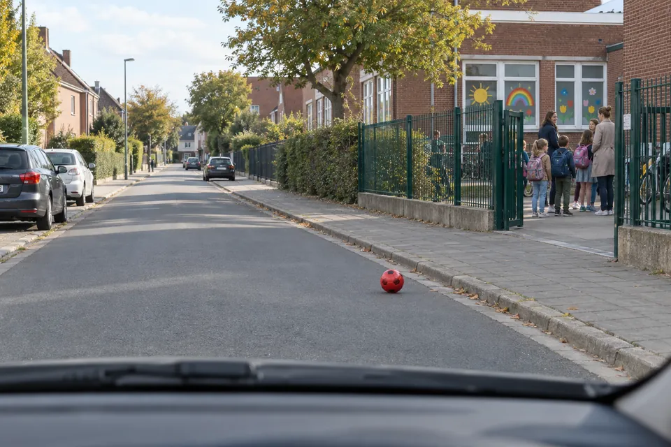 Balle rouge sur la chaussée près d’une école, avec des enfants et des adultes à l’entrée hors de la chaussée.